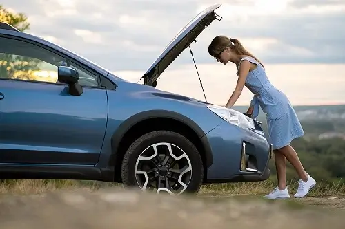 Common Auto Repair Issues Drivers Face in Gilbert, AZ | Ace Performance Auto. Image of a stranded young woman driver standing near her broken blue car with popped up bonnet, inspecting her car for battery problem or overheating engine.
